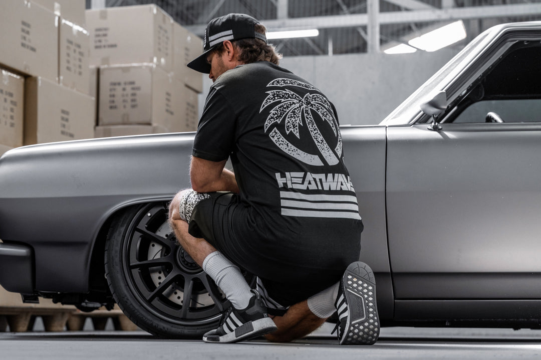 Man kneeling on the ground in front of a car wearing the Heat Wave Alloy T-Shirt.