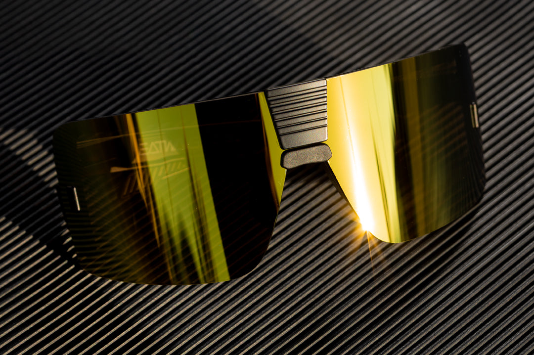 Sitting on top of a black table is the Heat Wave Visual Vector Sunglasses with black frame and gold lens.