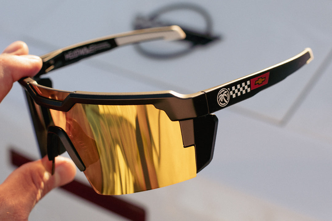 Hand holding the Heat Wave Visual Future Tech Sunglasses with black frame, corvette print arms and gold lens.