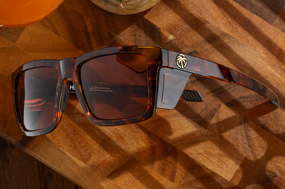 Lying on a wooden table is the Heat Wave Visual Performance Vise Sunglasses with tortoise frame, brown lenses and matching side shields.