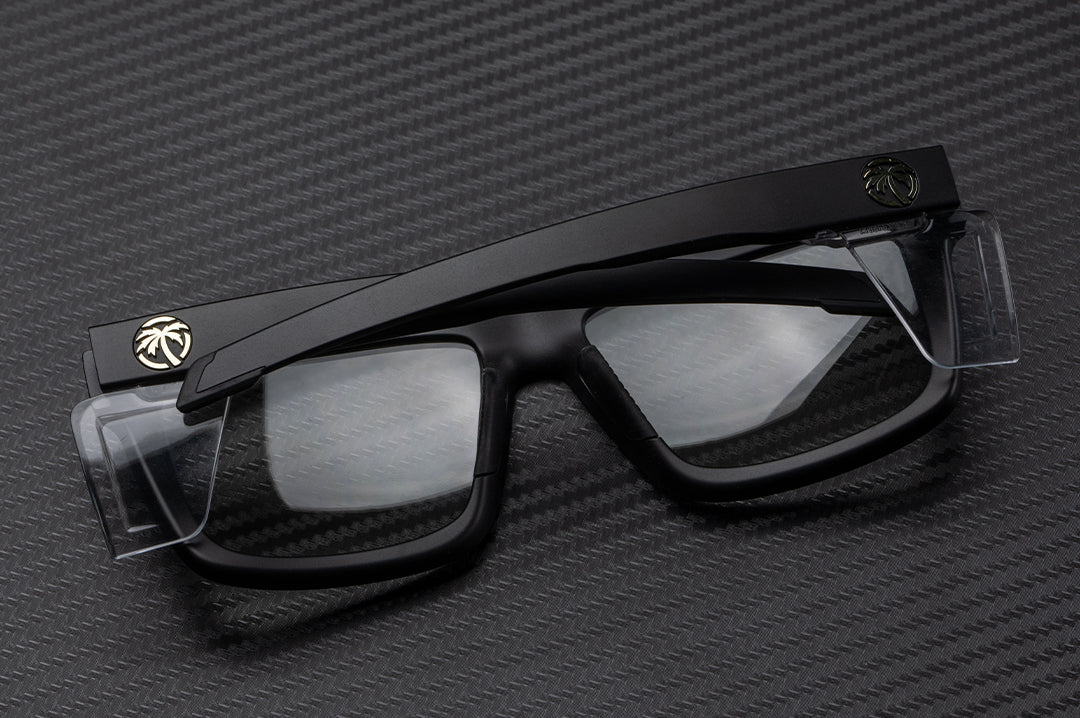 Laying on a table is the Heat Wave Visual Performance Vise Sunglasses with black frame, clear lenses and clear side shields.