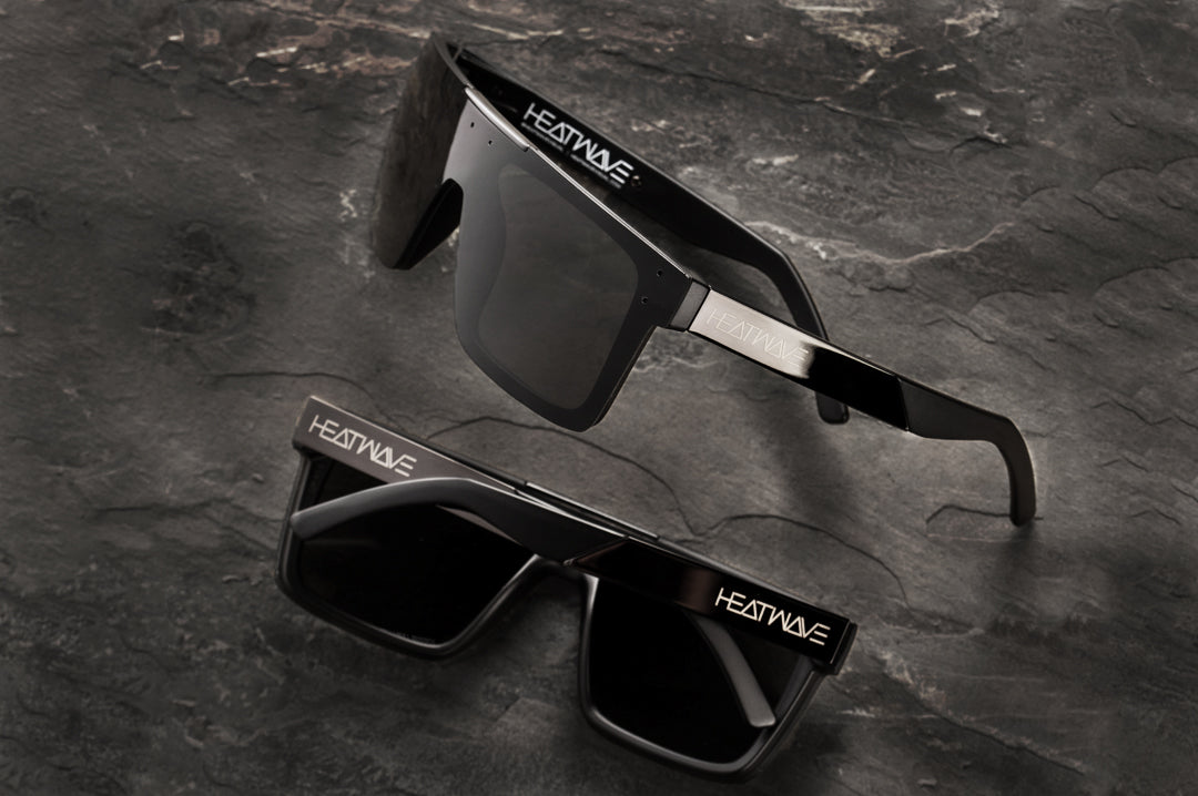 Two Heat Wave Visual Quatro Sunglasses with black frame, black metal arms and black lens lying on concrete.