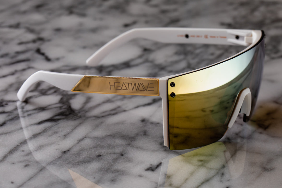 Side view of Heat Wave Visual Lazer Face Sunglasses with white frame, gold metal arms and gold lens on a marble counter.