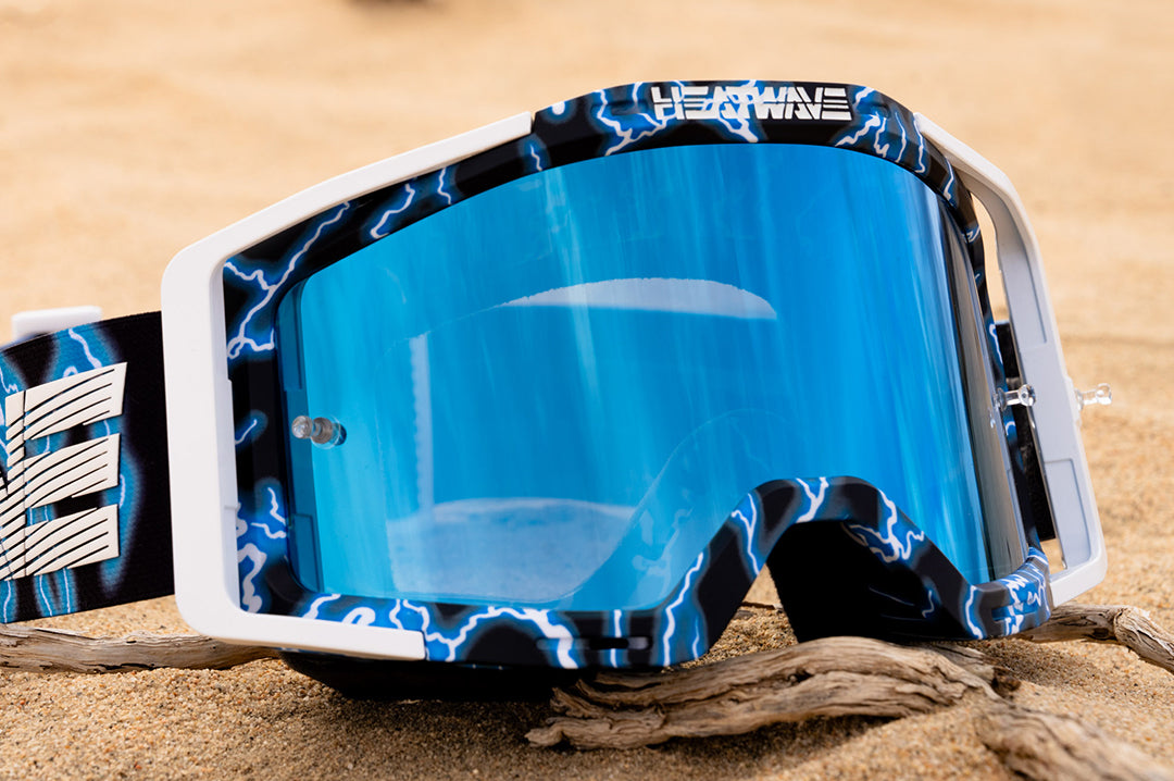 Close up shot of the Heat Wave Visual MXG 250 Motosport Goggle in the Lightning color way with a blue lens on the sandy ground in the desert.