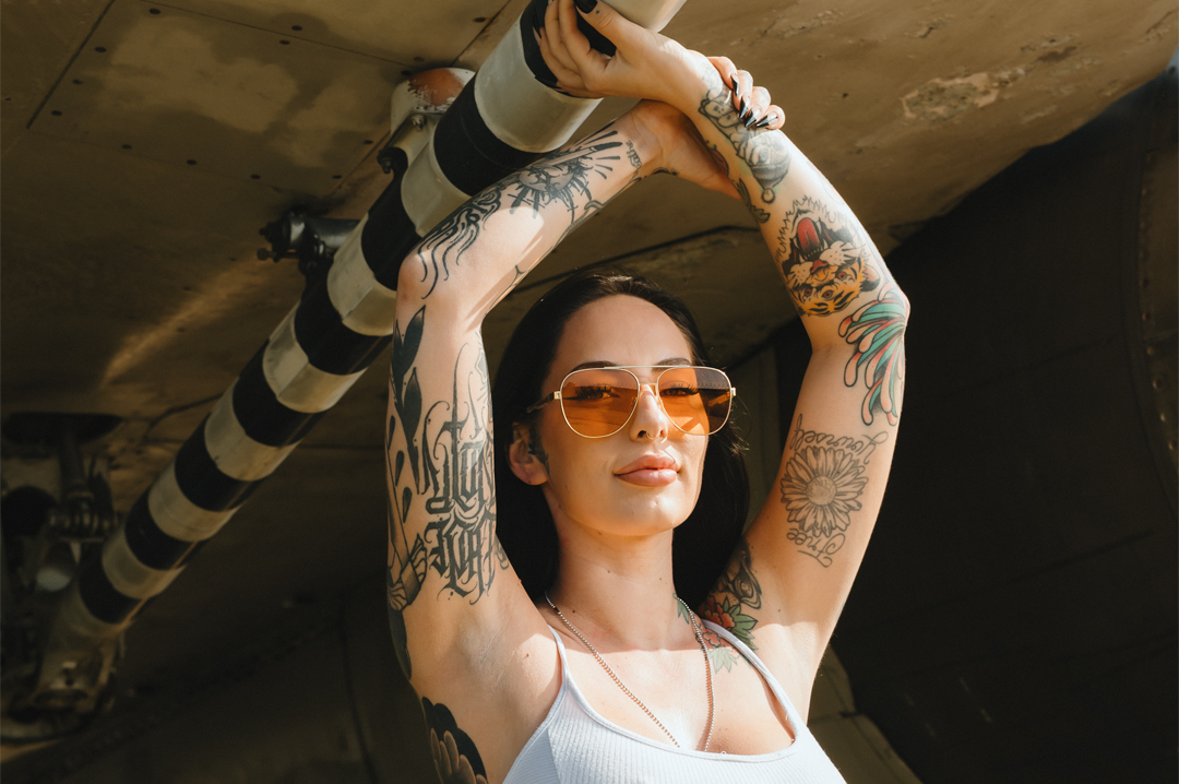Woman with tattoos wearing Aviator sunglasses with a polished gold frame and amber lenses under an F-14 airplane.