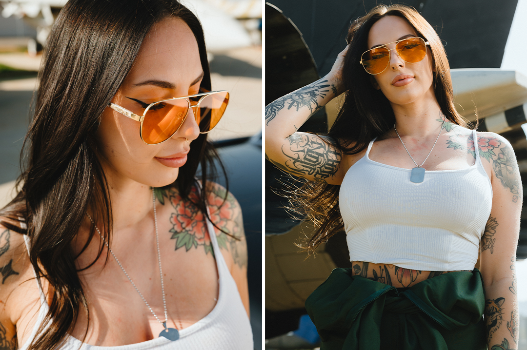 Woman wearing aviator sunglasses with a polished gold frame and amber lenses, paired with a white top and visible tattoos.