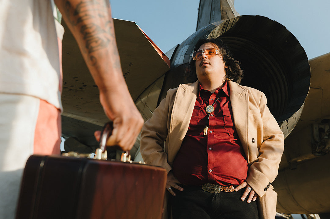 Person in a red shirt and beige coat and Polished Gold frame and amber lenses aviator sunglasses standing next to a F-14 airplane, holding a suitcase.