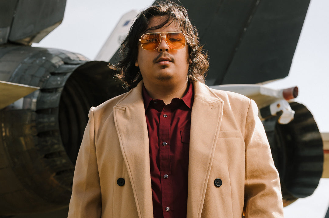 Person wearing a beige coat and Polished gold frame and amber lenses sunglasses in front of an F-14 airplane