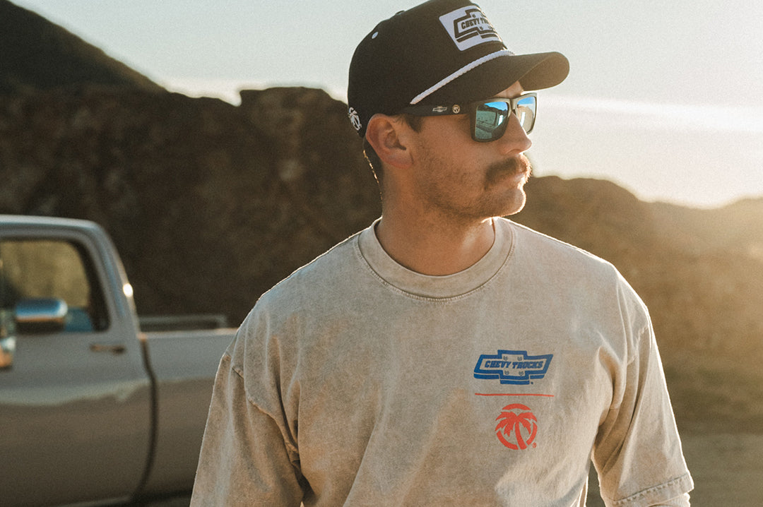 Man wearing a Chevy Truck and Heat Wave Visual XL Vise Sunglasses with black frame, Chevy Truck print arms and Galaxy lens standing in front of a truck with mountains in the background