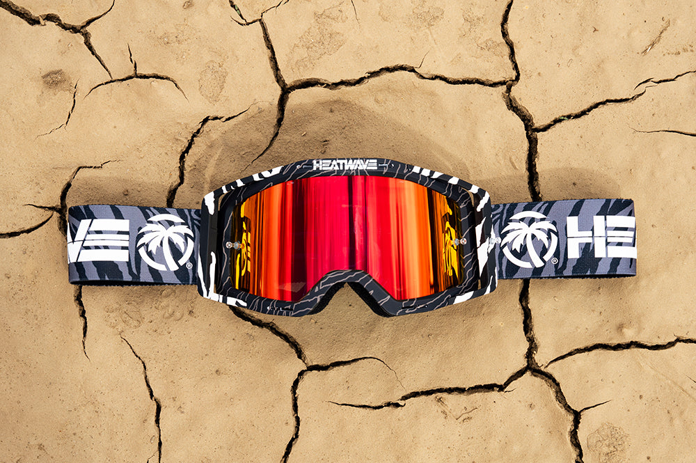 Lying on dirt is the Heat Wave Visual MXG 250 Motorsport Goggles in the Patrick Evans zebra stirpe color with firestorm red lens.