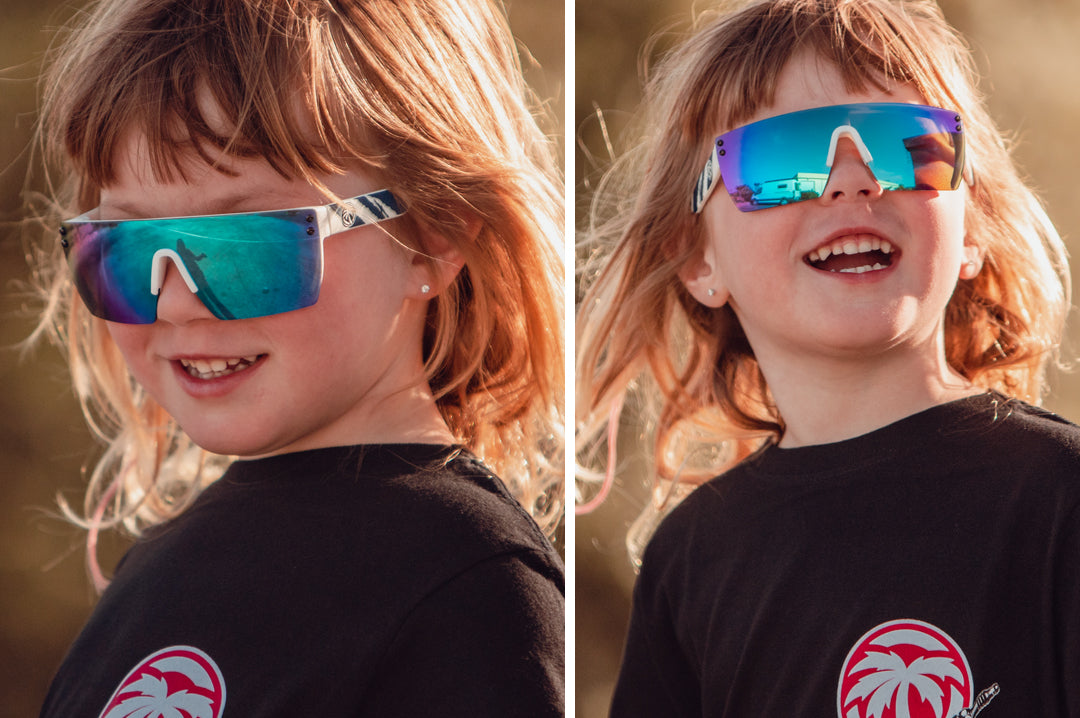 Smiling toddler girl wearing Heat Wave Visual Lazer Face kids sunglasses white frame, red white and blue print arms and galaxy blue lens.