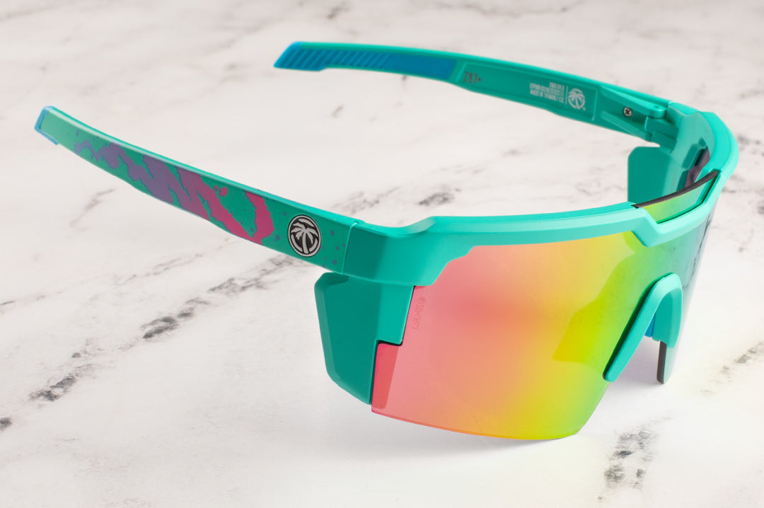 Sitting on a marble counter is the Heat Wave Visual Future Tech Sunglasses with teal frame, hypersplash print arms and spectrum pink yellow lens. 