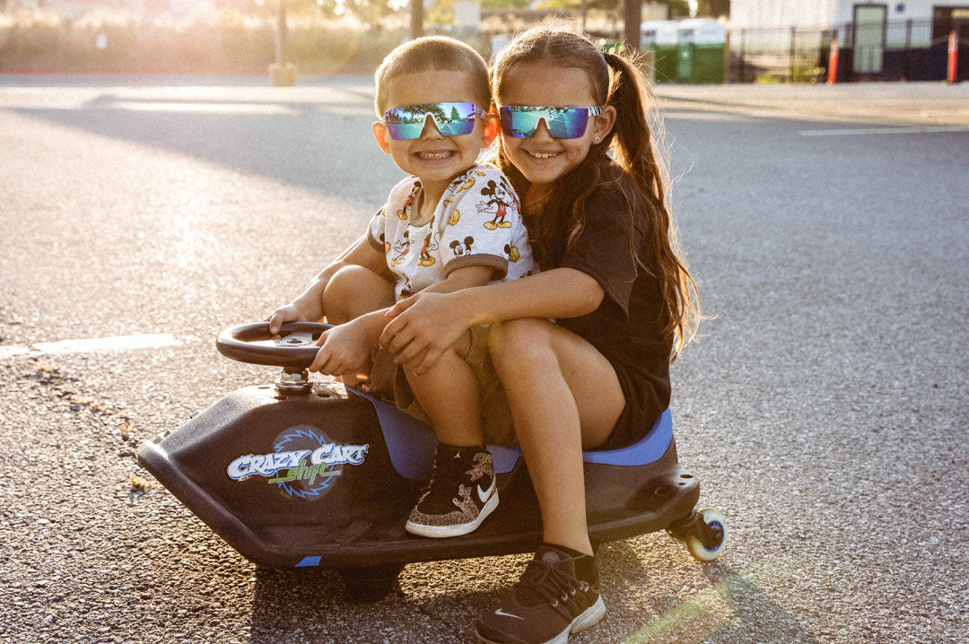 Young Siblings riding a crazy cart wearing the Heat Wave Visual Lazer Face kids sunglasses white frame, USA print arms and galaxy blue lens.