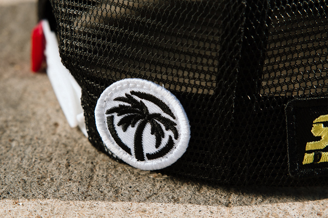 Close up shot of the embroidered palm tree logo on the backside of the Little Trouble X Heat Wave Youth Trucker Hat on a concrete floor.