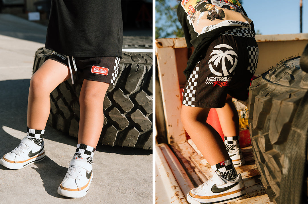 Front and backside photos of a young boy wearing the Little Trouble X Heat Wave Kids Board Shorts outside next to a big tire on the ground.