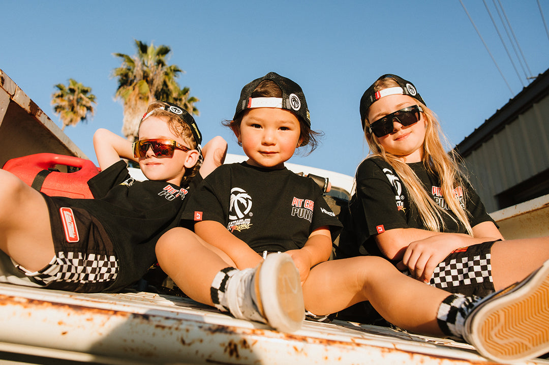 Three young kids sitting on the back of a truck wearing the Front and backside of the Little Trouble X Heat Wave Kids Board Shorts.