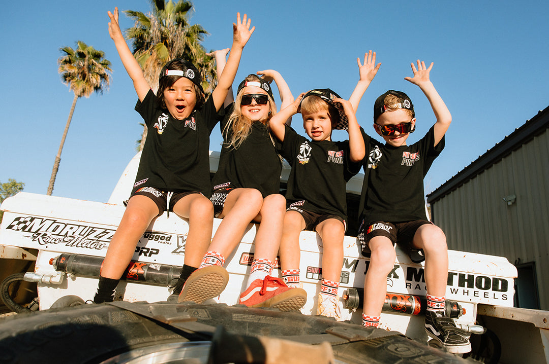 Four young kids sitting on the back of a truck wearing the Little Trouble X Heat Wave Kids T-Shirt with the hands raised up in the air.