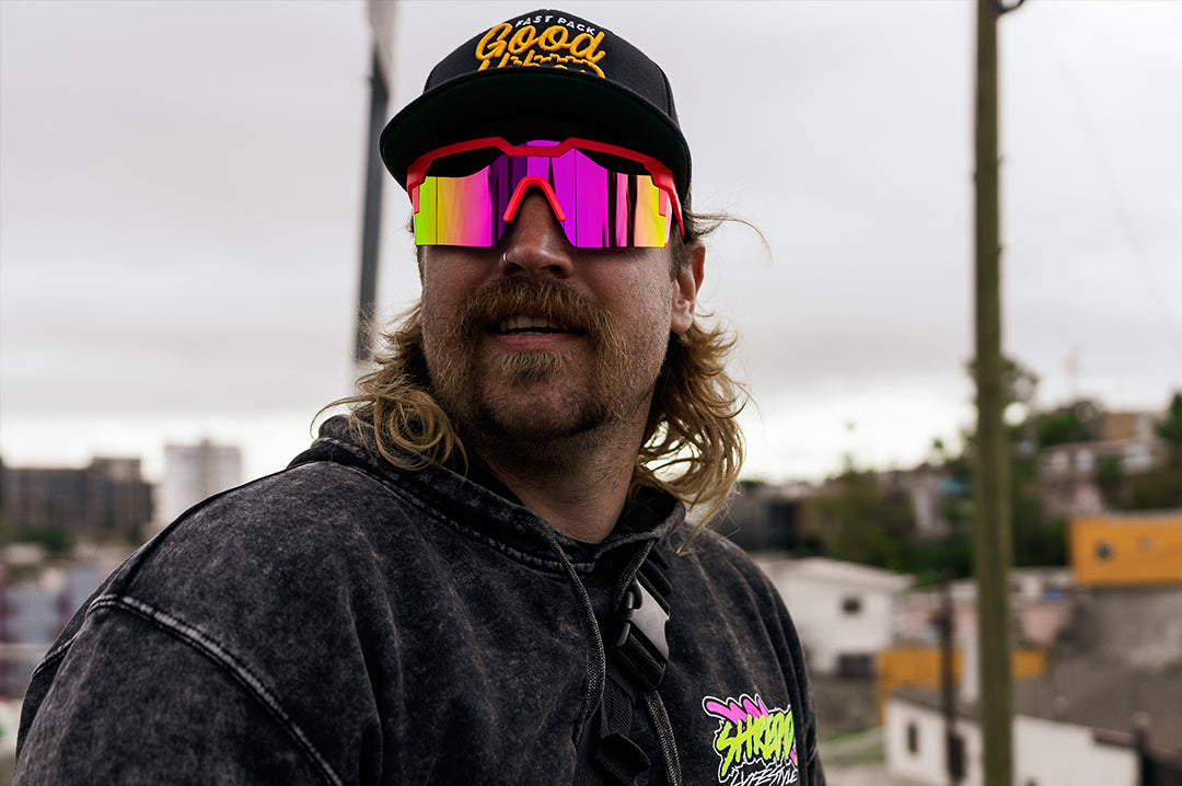 Man with a mullet wearing Heat Wave Visual Future Tech Sunglasses with pink frames, standup arms and spectrum pink yellow lens.
