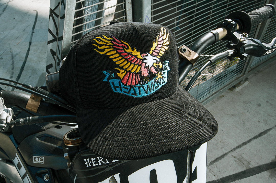 the Heat Wave x Tall Boy Eagle Cord Hat - Black on top of a motorcycle