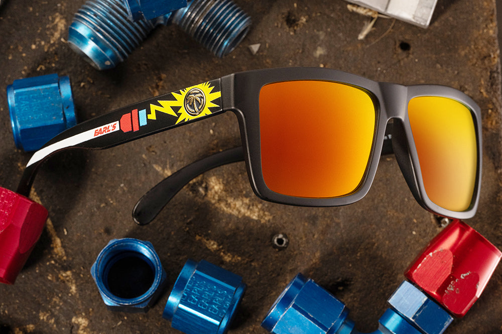 Lying on a the ground is the Heat Wave Visual XL Vise Sunglasses with black frame, Earl's x HWV collab and sunblast orange yellow lenses.