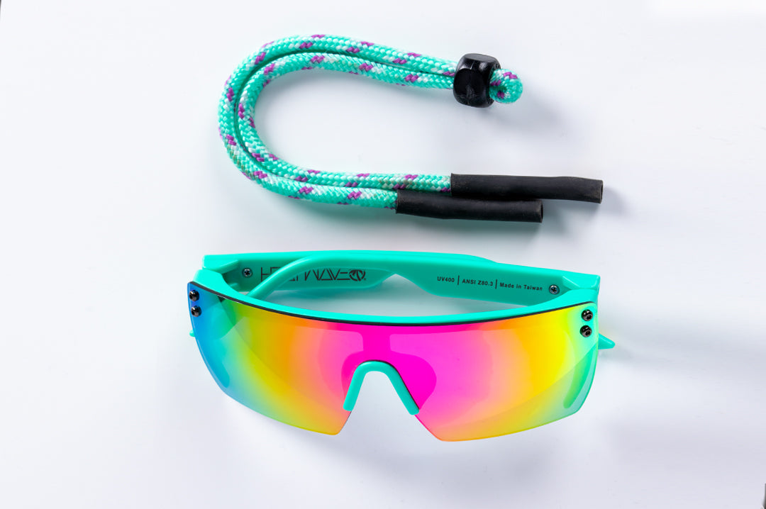 Heat Wave Visual Lazer Face Kids Sunglasses with teal frame, brush print arms and spectrum pink yellow lens sitting on a table.
