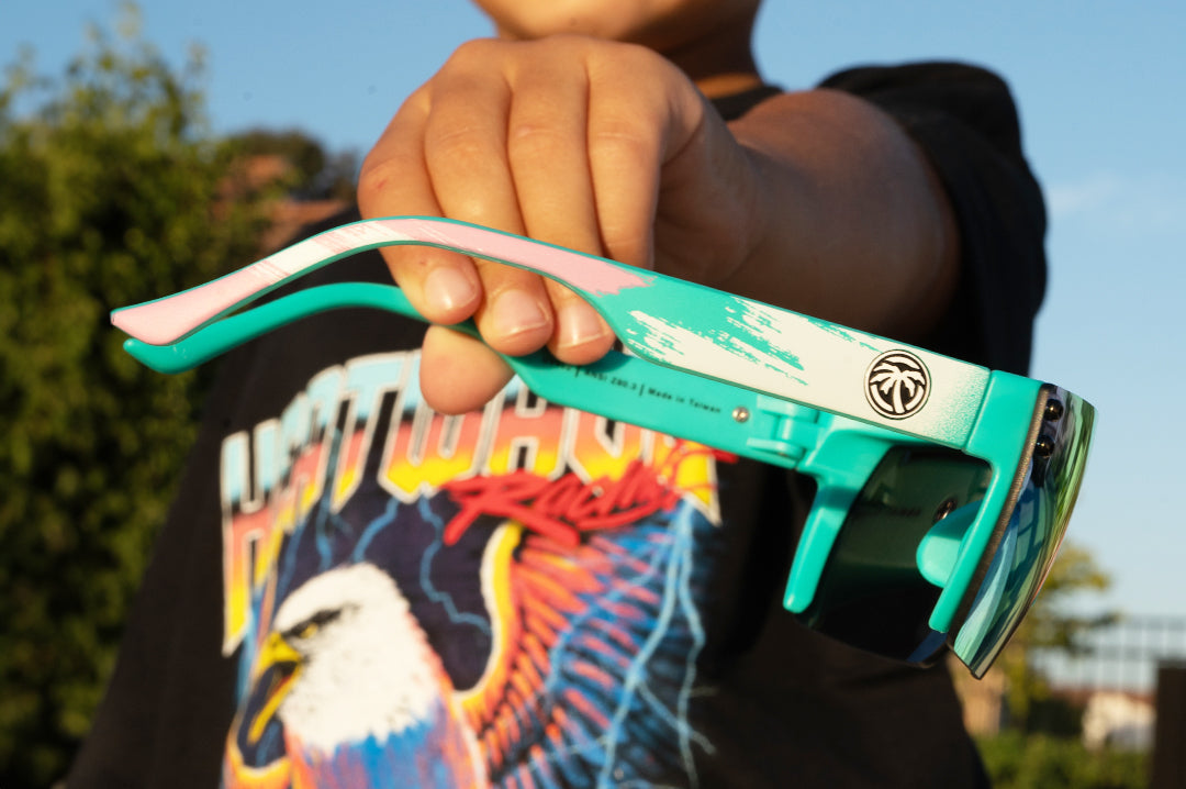 Kid holding up a pair of Heat Wave Visual Lazer Face Kids Sunglasses with teal frame, brush print arms and spectrum pink yellow lens.
