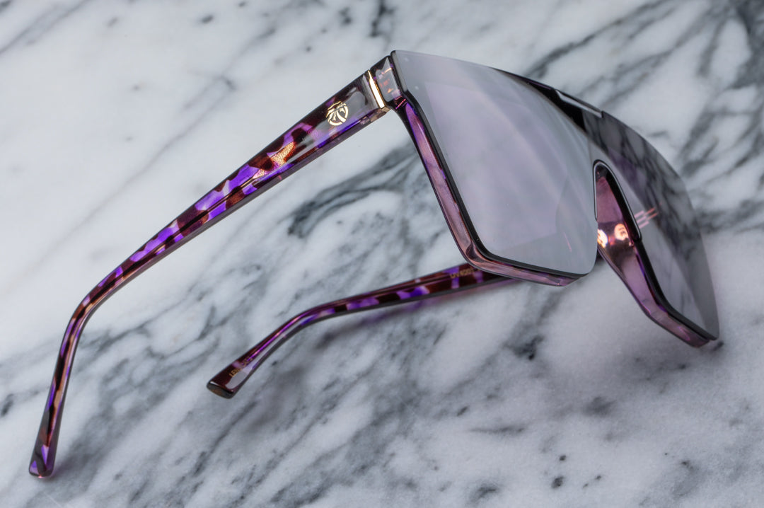Laying on its side is the Heat Wave Visual Women's Clarity Sunglasses with velvet tortoise frame and purple lens.