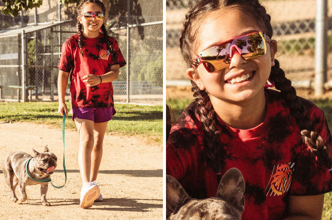 Young girl walking her dog wearing the Heat Wave Visual Lazer Face kids sunglasses pink frame, pink splatter print arms and rose gold lens.