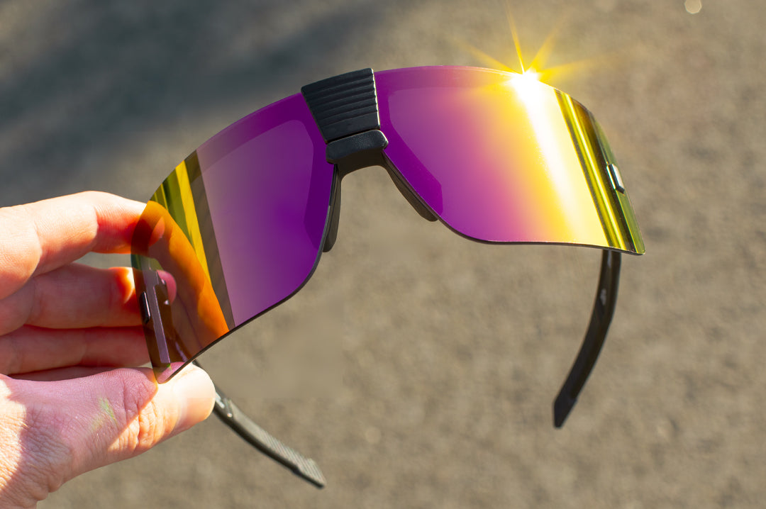 Heat Wave Visual Vector Sunglasses with black frame and spectrum pink yellow lens being held up by a hand outside.