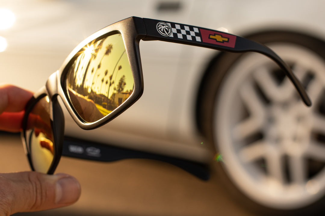 Hand holding up the Heat Wave Visual XL Vise Sunglasses with black frame, corvette print arms and gold lenses.