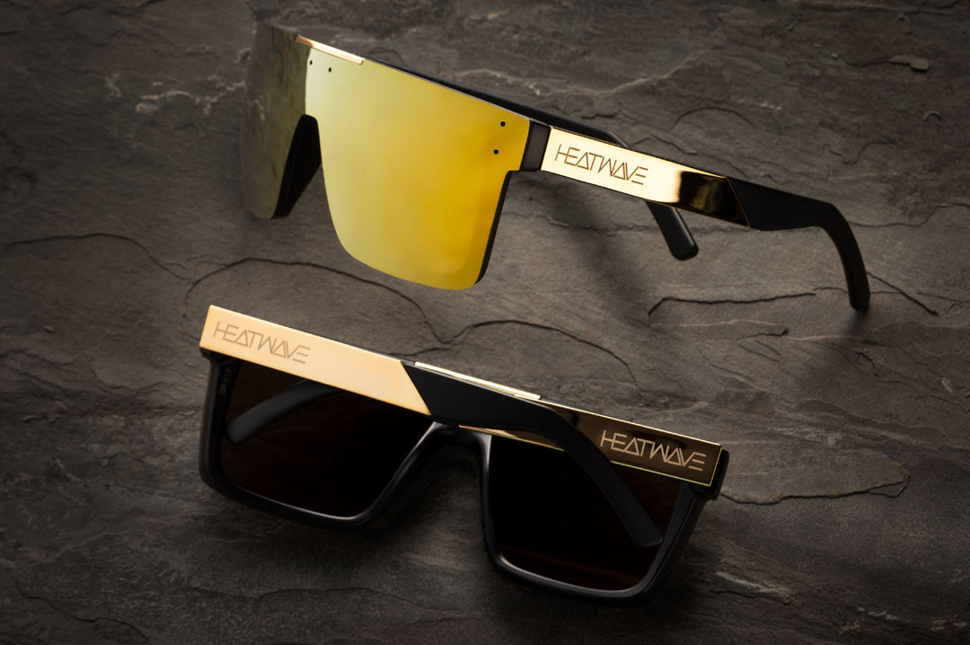 Two pairs of Heat Wave Visual Quatro Sunglasses with black frame, gold metal arms and gold lens lying on a slab of rock. 