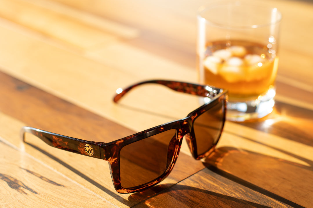 Heat Wave Visual Tortoise XL Vise Sunglasses sitting on a wooden floor next to a glass of whiskey.
